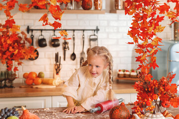 Cute little girl happily rolling out dough with a red rolling pin while baking in a rustic kitchen richly decorated with vibrant orange and red fall leaves. autumn season,  autumn concept,  fall