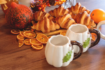 Cozy autumn breakfast setting with fresh croissants, dried orange slices, decorative gourds, and two ceramic pumpkin-shaped mugs on a wooden table. autumn season,  autumn concept,  fall