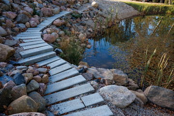 A stone pathway runs next to a pond bordered by rocks and reeds. The pond reflects vegetation and sky, with scattered leaves on its surface.