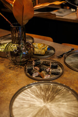 A table is arranged with metallic patterned plates, sushi rolls, a pastry, and a glass vase with dried leaves under warm lighting and earthy tones.