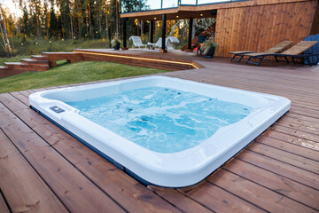 Modern hot tub set into a polished wooden deck with bubbling water, lounge chairs, string lights, and a wooden facade structure surrounded by tall trees.