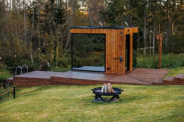 A modern wooden sauna with glass panels sits on a raised deck with string lights, a fire pit, a water feature, and surrounded by grassy areas and trees.