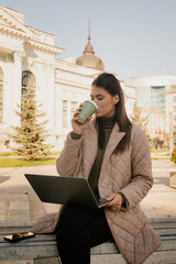 Professional woman uses laptop outside, enjoying coffee in an urban plaza with historic architecture.