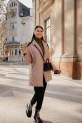 Stylish woman in a pink quilted coat strolls along a sunny city street, carrying coffee and phone.