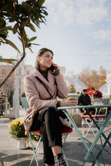 Casual street scene: a woman in a pink quilted coat chats on her phone at an outdoor cafe, holding a coffee.