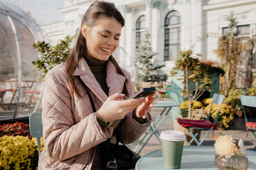 A cheerful woman sits at an outdoor cafe, texting on her phone with a takeout coffee nearby.