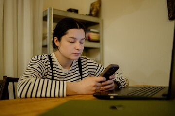 A young woman with a cell phone in hand and a laptop sits at the dining room table in her kitchen in the night light, working online
