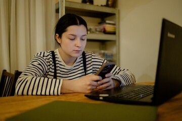 A young woman with a cell phone in hand and a laptop sits at the dining room table in her kitchen in the night light, working online