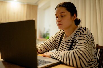 A young woman with a cell phone in hand and a laptop sits at the dining room table in her kitchen in the night light, working online