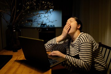 A woman sitting at a table with a laptop, yawning. She is wearing a striped shirt and has her hair tied back. The background shows a kitchen area with soft lighting.