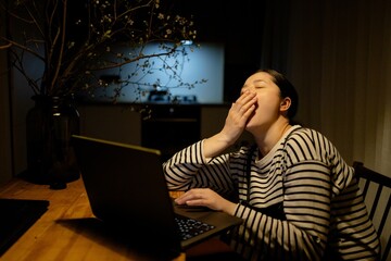 A woman sitting at a table with a laptop, yawning. She is wearing a striped shirt and has her hair tied back. The background shows a kitchen area with soft lighting.