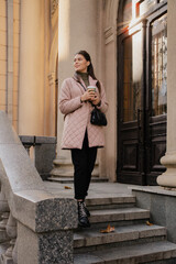 Stylish woman stands on stone steps holding a coffee, wearing a pink quilted coat and black boots in autumn.