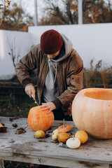Stylish man carving halloween pumpkin on rustic wooden table in autumn garden. Fall harvest....