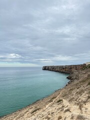 Rocky coastal cliffs meet calm turquoise sea under a cloudy sky, creating a dramatic seascape.
