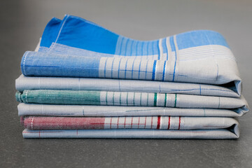 Stack of three neatly folded handkerchiefs in various colors (blue, green, and red) with grey centers and a plaid pattern, photographed on a solid gray surface.