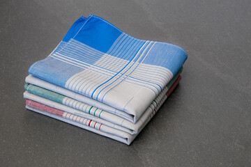 Stack of three neatly folded handkerchiefs in various colors (blue, green, and red) with grey centers and a plaid pattern, photographed on a solid gray surface.