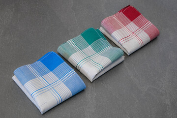 Set of three neatly folded handkerchiefs in various colors (blue, green, and red) with grey centers and a plaid pattern, photographed on a solid gray surface.