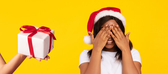 African American Man In Santa Hat Holding Christmas Gift Surprising Woman While She Covering Eyes Standing Over Yellow Studio Background. Happy New Year, Xmas Presents Delivery Concept