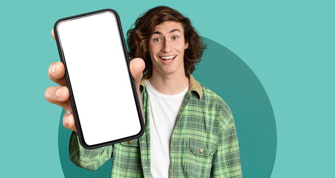 A young man with curly hair is smiling and showing a blank smartphone screen. He wears a green plaid shirt and is set against a bright teal backdrop.