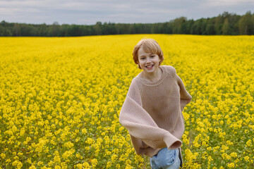 Fototapeta premium Smiling Caucasian girl, about 9 years old, front view, wearing oversized beige sweater, standing in yellow flower field, look at camera. Concept of childhood, joy, countryside, freedom.