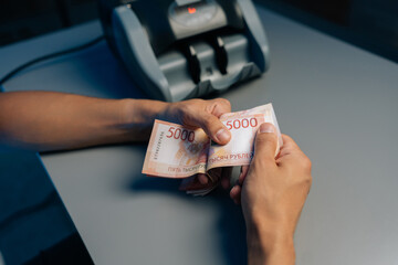Close-up cropped shot of cashier male counting Russian rubles banknotes late at night after closing store using money counting machine in background. Concept of financial accounting in modern office.