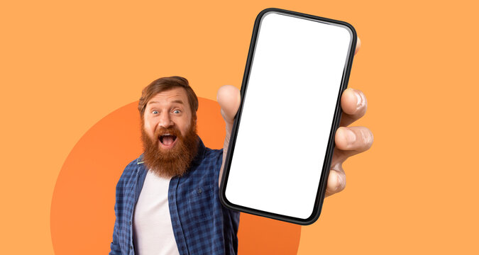 A young man with a bushy beard and cheerful expression is holding up a smartphone with a blank screen. He is wearing a plaid shirt and standing against a vibrant orange backdrop. - Powered by Adobe