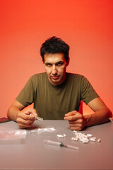 Vertical portrait of serious man preparing to snort cocaine with rolled banknote sitting at table with pills and syringe on red background, representing drug addiction and substance abuse.