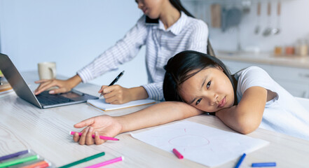 Young korean mother working from home, ignoring her offended little daughter, girl feeling lonely and abandonned while her asian mom busy with laptop computer
