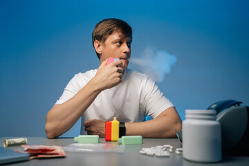 Drug dealer male smoking heated tobacco from sleek modern heat-not-burn tobacco device sitting at desk with money, pills and various drugs in front of him and exhaling smoke on blue background.