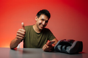 Successful cashier giving thumbs up while counting banknotes with money counter machine sitting at gay table, smiling looking at camera, radiating positivity and success, on isolated red background.