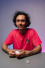 Vertical portrait of handsome young adult man with curly hair and pink t-shirt cutting fingernails using nail clipper sitting at gray table against purple background, smiling looking at camera.