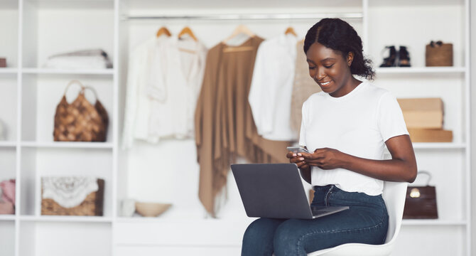 Happy African American Lady Using Laptop And Smartphone Browsing Internet Sitting In Wardrobe Among Clothes Shopping Online At Home. Internet Technology And E-Commerce Concept - Powered by Adobe