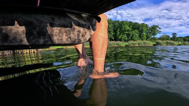 Low angle perspective of a man's legs dangling from a wooden pier, dipping his feet and splashing in the refreshing lake water during a beautiful sunny day, creating ripples and reflections