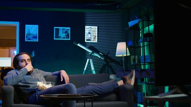 Homeowner bored at home munching on snacks, using TV to binge series on subscription based streaming services. Man sitting on couch, wasting time with VOD shows on TV, eating popcorn, camera B