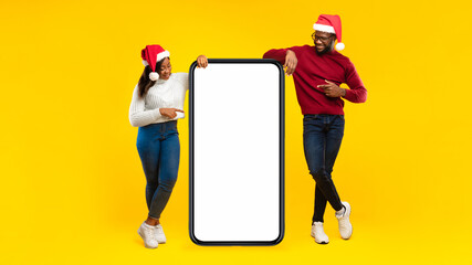Great Application For Smartphone. African Millennial Couple Wearing Santa Hats Pointing Fingers At Big Mobile Phone With Blank Screen Advertising Great Xmas Offer On Yellow Background In Studio