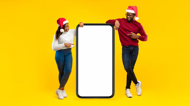 Great Application For Smartphone. African Millennial Couple Wearing Santa Hats Pointing Fingers At Big Mobile Phone With Blank Screen Advertising Great Xmas Offer On Yellow Background In Studio - Powered by Adobe