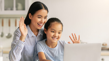 Happy asian mother and daughter having video call, waving and smiling at laptop screen, sitting in...