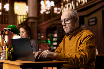Intellectual mature person reviewing academic text on a laptop at the university, sitting in...