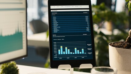 Fototapeta premium Modern workspace with analytical data paperwork documents in bright office environment with potted plants. Tablet on desk with digital statistics charts displayed on screen, close up shot