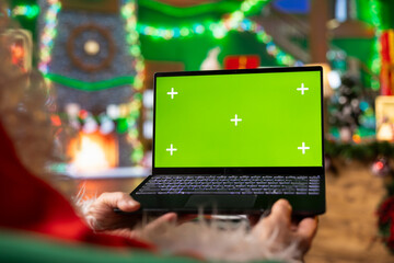 Santa using green screen laptop in workshop to check heartfelt messages from kids worldwide. Father Christmas reviews wishes sent by children via email using mockup notebook