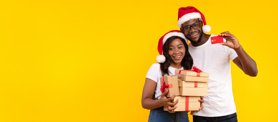 Happy African American Couple Showing Credit Card Holding Christmas Gifts Boxes Wearing Santa Hats Standing Over Yellow Background, Smiling To Camera. Xmas Presents Shopping Concept