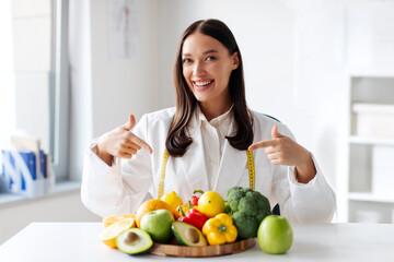 Portrait of professional female dietitian pointing at fresh fruits and vegetables on table and smiling. Weight loss consultant recommending healthy eating