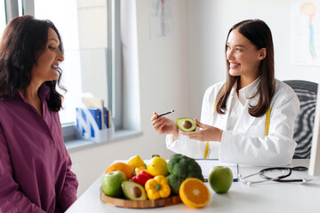 Cheerful woman dietician giving advices on improving diet, holding fresh avocado in hand and talking with mature female patient