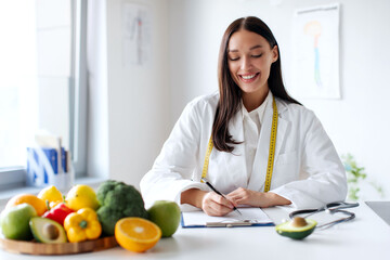 Dietitian making balanced corrective diet plan for patient, writing in clipboard, sitting at office desk with raw vegetables and fruits, free space