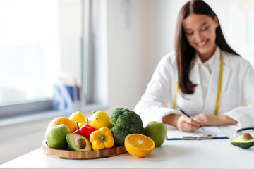 Focus on wooden board with raw fruits and vegetables on table with young woman nutritionist working on foreground writing diet plan for patient