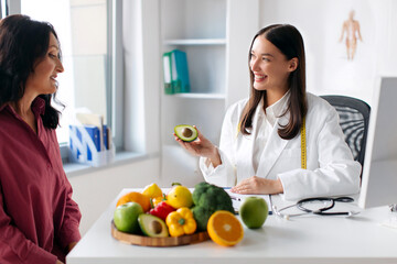 Happy lady nutritionist telling mature woman patient about benefits of eating vegetables, having consultation, female food expert holding avocado in her hand and speaks
