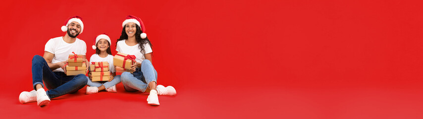 Christmas Presents. Arab Parents And Little Daughter Holding Xmas Gifts Boxes Wearing Santa Hats Sitting In Studio Over Yellow Background. Holidays Season And Winter Offers Concept