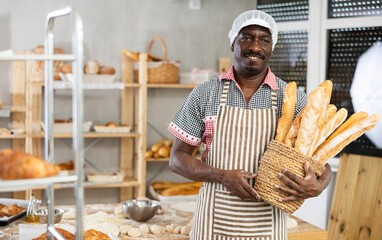 Male baker with basket of baked goods behind bakery counter inviting to buy fresh bread