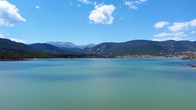 panoramic view of the tranquil Lake Doxa in Greece, nestled amidst lush green mountains under a vibrant blue sky with fluffy white clouds. Ideal for showcasing natural beauty, travel, and peaceful esc