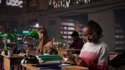 Black girl reading books and handwritten class notes right before the exam, making preparations all night to get a high grade for scholarship. Young adult shows dedication and ambition. Camera B.
